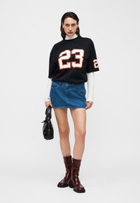 Femme portant une chemise noire oversize avec le numéro blanc 23, un haut blanc à manches longues, une jupe en denim bleu, des bottes bordeaux, tenant un sac à main noir.