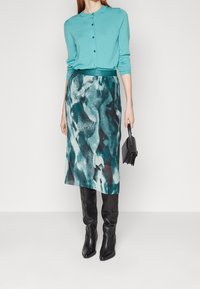 Cardigan teal avec fermeture à boutons associé à une jupe genou à motifs teal et noir. Le modèle porte des bottes noires montantes.