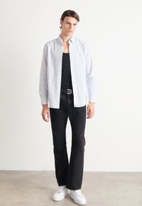 Camisa de botones de rayas azul claro sobre una camiseta negra, combinada con jeans negros y zapatillas blancas. Diseño simple y moderno con un corte relajado.