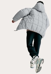 Person iført en lys grå puffjakke, sorte bukser, hvide sokker og sneakers, der går med det ene ben løftet foran en ensartet baggrund.