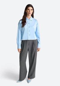 Himmelblaues, tailliertes Hemd mit langen Ärmeln, kombiniert mit grauen, weit geschnittenen Hosen und himmelblauen spitzen Schuhen.