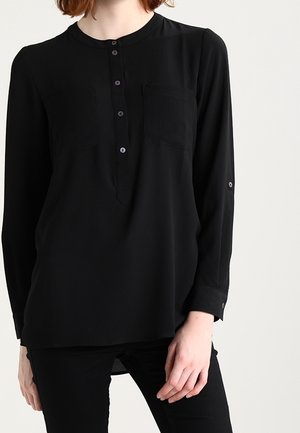 Femme portant un chemisier noir à manches longues avec une patte de boutonnage sur le devant et une poche poitrine, associé à un pantalon noir.