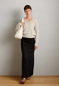 Žena s krátkými vlasy má na sobě krémový pletený svetr, černou maxi sukni, sandály a nese velkou bílou kabelku přes rameno u šedé zdi.