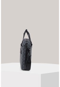 Czarna torba tekstylna z wzorem monogramu, cienkimi uchwytami, zapięciem na zamek oraz odpinanym paskiem. Widok z boku podkreślający prostokątny kształt i fakturę.