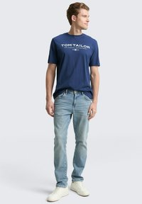 Marineblauwe T-shirt met wit "TOM TAILOR" logo, gedragen met lichtblauwe spijkerbroek en witte sneakers. Model poseert met de gezicht naar links.