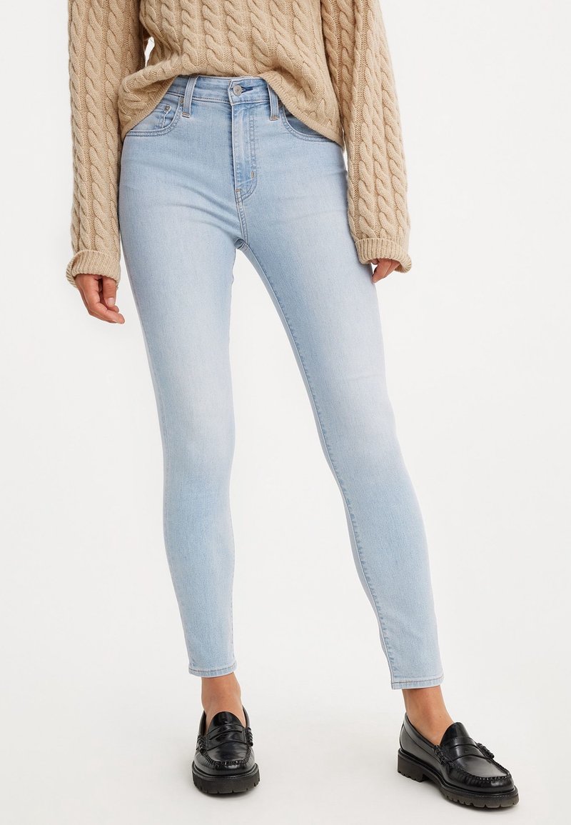 Personne portant un jean skinny bleu clair, un pull beige en maille torsadée et des mocassins noirs, debout devant un fond blanc uni.