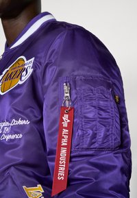 Fialová bomber bunda s logom Los Angeles Lakers, rebrovaným golierom, zipsovým vreckom a červeným štítkom s textom "ALPHA INDUSTRIES". Saténový povrch.