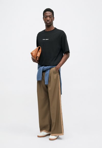 Schwarzes T-Shirt mit weißem Logo, blauer Pullover um die Taille gebunden, braune weite Hose mit beigen Seitestreifen und cremefarbene Schuhe. Hält eine braune Clutch.