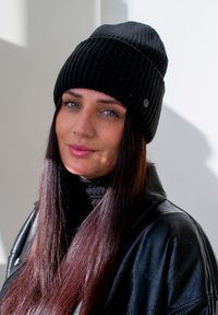 Berretto a coste nero indossato con un dolcevita nero e una giacca di pelle. Capelli lunghi e lisci in vista. La luce soffusa crea ombre sottili.