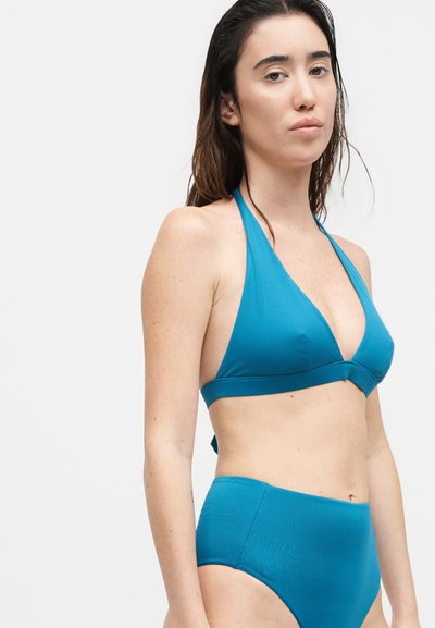 Γυναίκα με μακριά σκούρα μαλλιά που φοράει τοπ μπικίνι χρώματος τιλ με halter και μπικίνι σλιπ με ψηλόμεση γραμμή σε απλό λευκό φόντο.