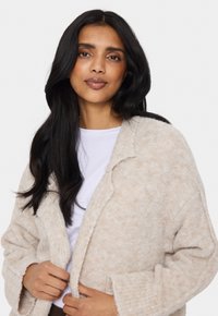 Ljusbeige oversized cardigan tillverkad av mjukt, strukturerat tyg, med vida ärmar och krage med notch. Bärs öppen över en vit skjorta.