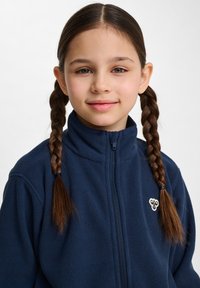 Mörkblå fleecejacka med hög krage och dragkedja, med en broderad logga på vänster sida. Slät textur, avslappnad passform.