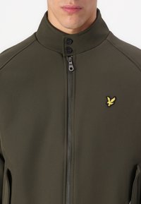 Giacca verde oliva con colletto alto, chiusura frontale con zip e due bottoni. Presenta un piccolo logo ricamato giallo sul petto.