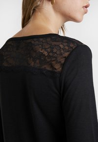 Svart långärmad topp med spetsöverdrag på axlarna, med en mjuk textur och rundad halsringning. Spetsen ger detalj längs kanten.