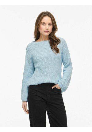 Woman wearing light blue knitted sweater with long sleeves and black pants, standing with one hand in pocket against white background.