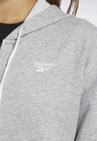 Felpa grigia con zip realizzata in tessuto morbido, caratterizzata da un logo Reebok bianco sul lato sinistro del petto, con un cappuccio grigio dalla texture in cotone e lacci.