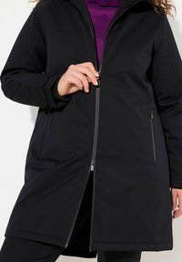 Svart zip-up jacka med huva, som har långa ärmar, två zip-fickor och en slät, vattenavvisande yta. Detaljer inkluderar ett kontrasterande innerfoder.