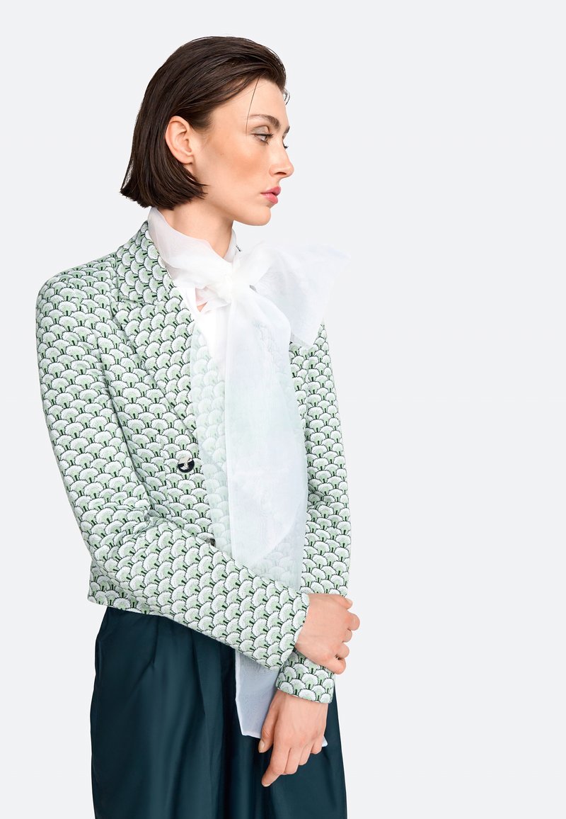 Sacou cu model în valuri verzi, design ajustat și mâneci lungi; asortat cu un eșarfă mare, albă, transparentă legată la gât.