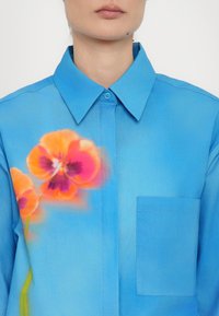 Light blue shirt with a collar, featuring a floral design in orange and pink on the left side, and a left chest pocket.