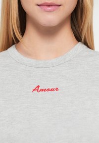 Γκρι μπλουζάκι με απαλή υφή, στρογγυλή λαιμόκοψη και κόκκινο κεντητό κείμενο «Amour» στο μπροστινό μέρος.