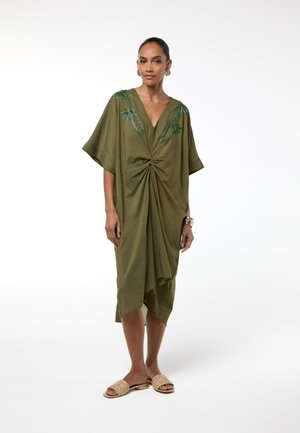 Mujer con vestido verde oliva hasta la rodilla con detalle de nudo y bordado de palmas, combinado con sandalias tejidas tipo slip-on y joyas de oro.