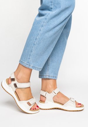 Pieds portant des sandales béiges à bout ouvert avec des sangles ajustables et du vernis à ongles rouge, associés à un jean court bleu clair.