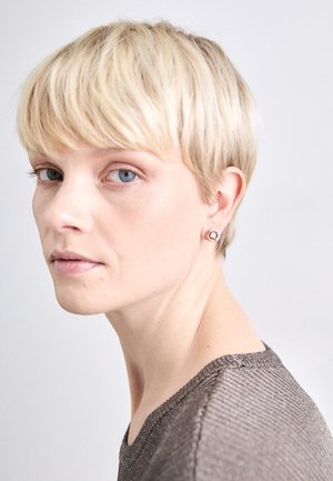 Cirkulære stud øreringe med en rosaguldfarve og en central klar sten, bæres af en person med kort blond hår og en struktureret grå top.