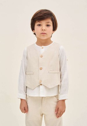 Beige Leinenweste mit Holzknöpfen, mit zwei frontalen Taschen, getragen über einem weißen Langarmshirt, kombiniert mit passenden beigen Hosen.