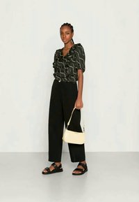 Blusa preta com detalhes em babados, padrão espiral branco, calças pretas de cintura alta e larga, sandálias pretas e uma mala cruzada amarelo claro.