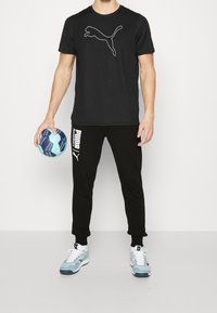 Sort T-shirt med hvid Puma-logo, sorte joggers med hvid tekst og blå og hvide sneakers. Holder en mønstret håndbold.