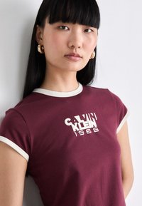 Burgundy short-sleeve t-shirt with white trim and logo saying "CALVIN KLEIN 1968." Soft fabric and fitted design.