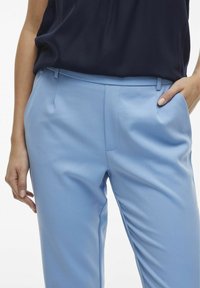 Pantalon bleu clair en tissu lisse ; doté de poches latérales et d'une coupe ajustée. Porté avec un haut sombre et des détails de plis subtils.