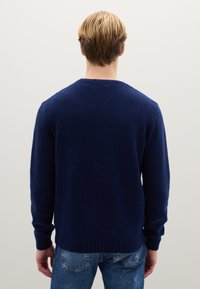 Marineblauer Strickpullover mit strukturiertem Muster, rundem Ausschnitt und gerippten Bündchen. Wird mit blauen Jeans aus Denim getragen, Ansicht von hinten.