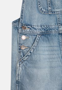 Lyseblå denimslipper med tre metallknapper foran, brystlomme og synlige sømdetaljer. Stoffet har en slitt tekstur.