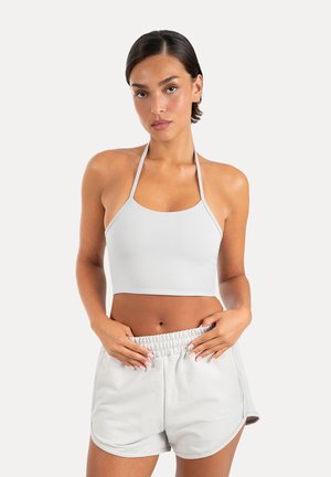 Kvinde med kort mørkt hår, iført en hvid halter crop top og lysegrå atletikshorts med elastisk talje mod en ensfarvet baggrund.