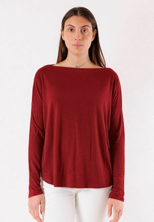 Donna con capelli lunghi e scuri che indossa una maglia bordeaux a maniche lunghe e pantaloni bianchi, in piedi davanti a uno sfondo bianco uniforme.