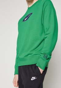Grön sweatshirt med långa ärmar, marinblå logotyp, rund halsringning och ribbstickade muddar. Matchas med svarta Nike sweatpants med vit logotyp.