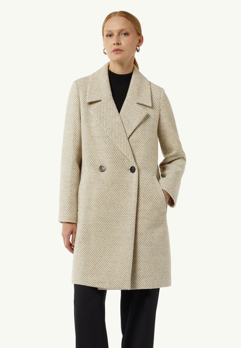 Beige visgraatjas met een dubbelbreasted ontwerp, brede revers en twee voorzakken. De stof heeft een gestructureerde uitstraling, knopen zijn zichtbaar.