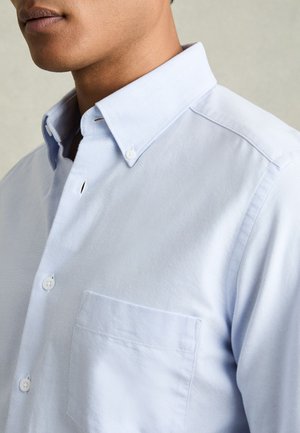 Lähivõte inimesest, kes kannab helesinist nööbitavat särki nööbitud kraega ja rinnataskuga, neutraalse tausta ees.