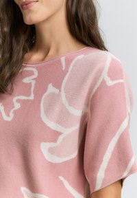 Růžový pletený svetr s bílým abstraktním vzorem, širokým výstřihem a krátkými rukávy. Měkká textura a uvolněný střih.