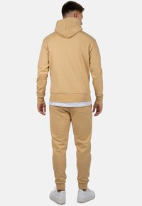 Uomo che indossa felpa con cappuccio beige e pantaloni da jogging abbinati con scarpe da ginnastica bianche, in piedi con la schiena rivolta verso chi guarda su sfondo semplice.