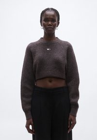 Pulover maro tricotat cu croială scurtă, cu model texturat, guler rotund și mâneci lungi, având un mic detaliu cu logo argintiu în față.