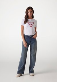 T-shirt bianca in cotone con logo triangolare rosso, abbinata a jeans in denim blu a gamba larga. La modella si trova in posizione con una mano sulla vita, le sneakers bianche sono visibili.