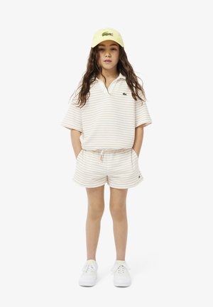 Polo a maniche corte a righe beige e crema con pantaloni coordinati, abbinato a un cappellino giallo. Caratterizzato da un piccolo logo verde. Sneakers bianche.