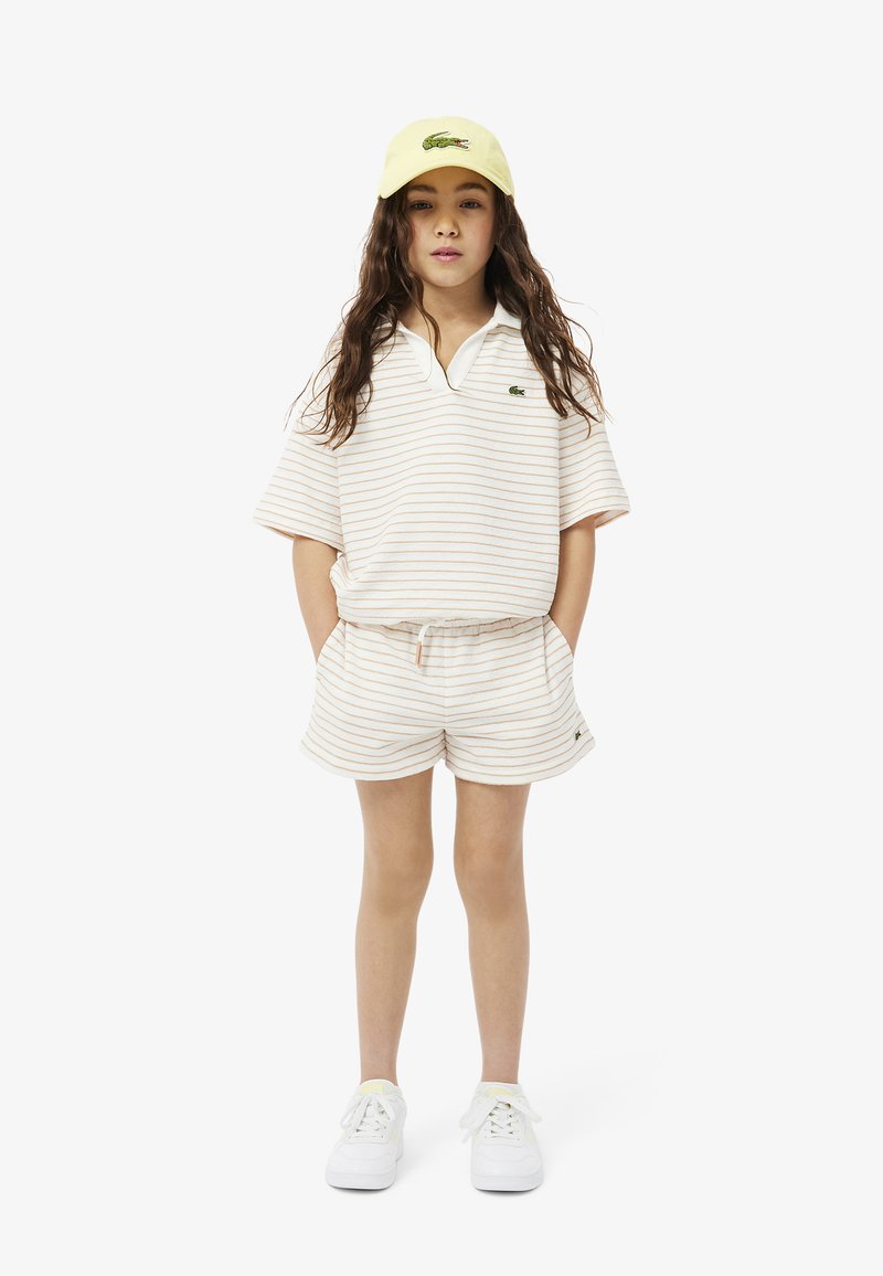 Polo de manga corta a rayas en beige y crema y pantalones cortos a juego, combinados con una gorra amarilla. Presenta un pequeño logo verde. Zapatillas blancas.