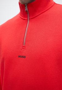 Pull rouge à fermeture éclair quart de zip avec zip argenté et logo noir "HUGO" centré sous le col sur le haut du torse d’une personne.