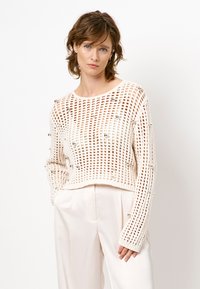 Vrouw met kort golvend haar, gekleed in een crème opengehaakte trui met zilveren kralen en bijpassende hoge taille wijde broek, staand tegen een witte achtergrond.