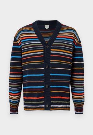 Muž stojaci s rukami v vreckách, oblečený v farebnom pruhovanom kardigáne, bielom tričku, tmavých nohaviciach a hnedých spoločenských topánkach na bielom pozadí.