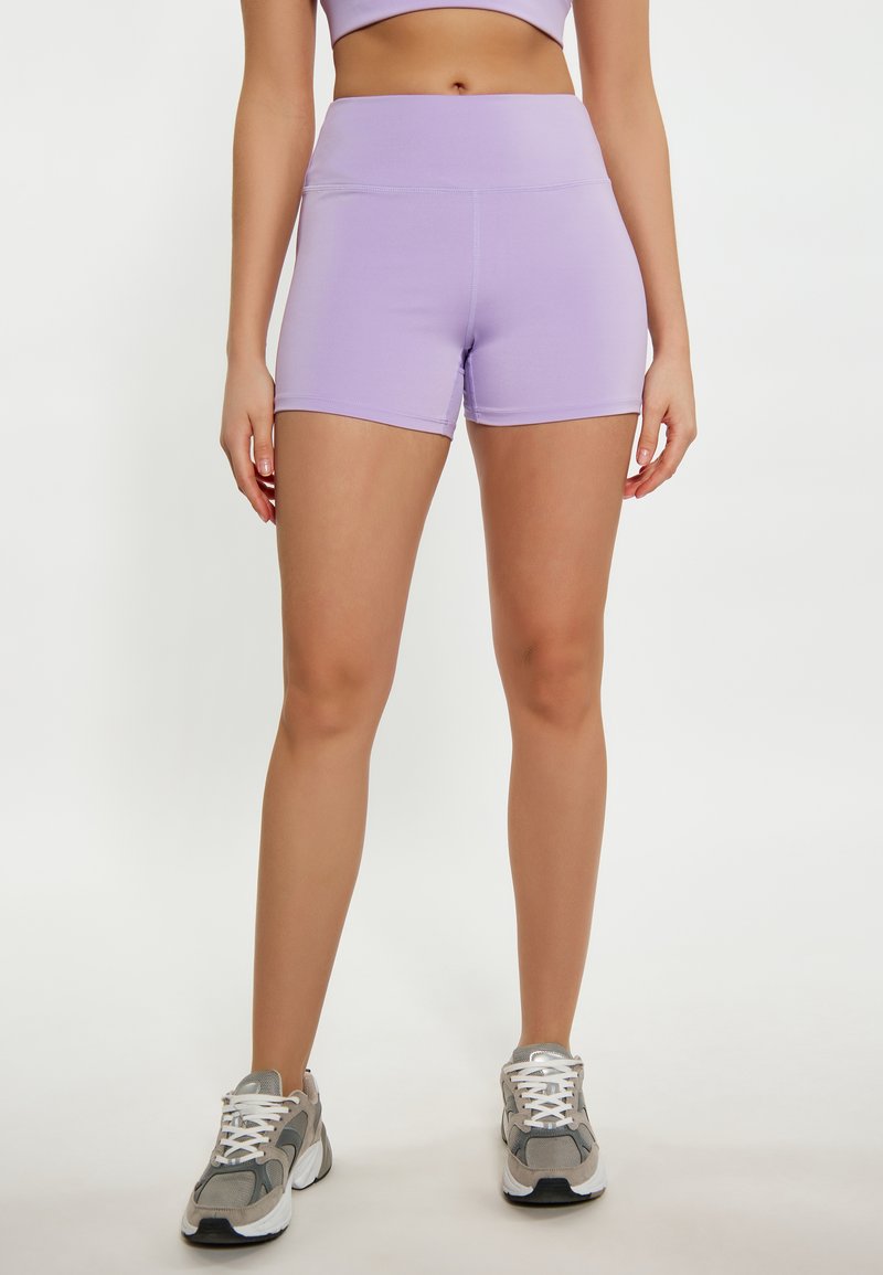 Shorts alti in lilac realizzati in tessuto elastico. Texture liscia e dettagli di cucitura minimi. Indossati con scarpe da ginnastica grigie e lacci bianchi.