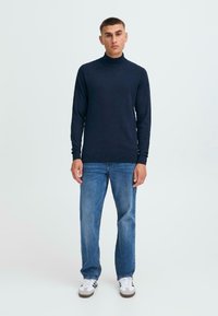 Pulover cu guler înalt de culoare bleumarin, într-un design ajustat, confecționat dintr-un material moale, asortat cu blugi de culoare albastru deschis și adidași albi.